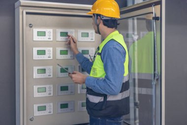Sert şapka ve güvenlik yeleği giymiş bir havalandırma teknisyeni birden fazla ekranı olan kontrol panelini kontrol ediyor. Teknisyen bir kalem kullanıyor ve elinde bir tablet tutuyor. Düzgün işlevsellik ve sistem verimliliği sağlıyor.