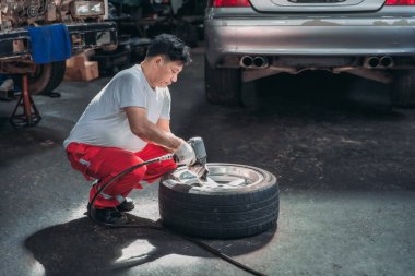 Garajda bir araba lastiği üzerinde çalışan yetenekli bir tamirci, pnömatik bir anahtar kullanıyor. Görüntü, otomotiv bakımı ve onarımı alanındaki adanmışlık ve teknik uzmanlığı gösteriyor.