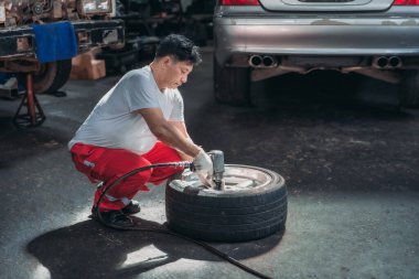 Garajda bir araba lastiği üzerinde çalışan yetenekli bir tamirci, pnömatik bir anahtar kullanıyor. Görüntü, otomotiv bakımı ve onarımı alanındaki adanmışlık ve teknik uzmanlığı gösteriyor.