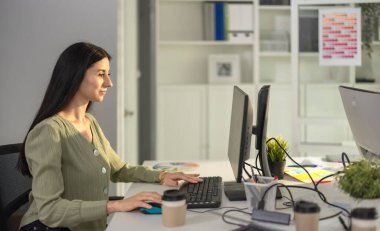 Profesyonel bir kadın modern ofiste masa başında oturur ve bilgisayarla uğraşır. Çalışma alanı ofis malzemeleri, kahve fincanları, saksı bitkileri ile organize edilir, verimli ve davetkar bir ortam yaratır.