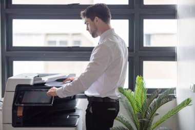 Profesyonel işadamı modern ofis ortamında fotokopi makinesi kullanıyor. Ofis görevlerinin verimliliği ve rutini, şirket ortamında belge yönetiminin önemini vurgulamak