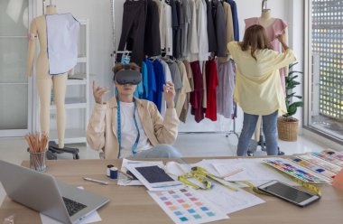 Modern bir stüdyoda moda ve kostüm tasarımcısı sanal tasarımları VR gözlükleri kullanarak araştırıyor, teknolojiyi yaratıcılıkla harmanlıyor, bir meslektaşı da arka planda bir elbise üzerinde çalışıyor..