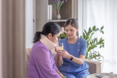 Mavi üniformalı özel bir bakıcı, rahat oturma odasında boyunluk takan bir kadına yardımcı olurken, bakıcı da hasta için ilaç hazırlıyor. Şefkat, destek ve profesyonel sağlık hizmetleri.