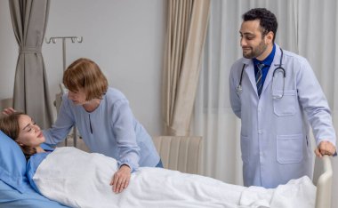 Bir doktor ve ilgili bir anne, hastane yatağında dinlenen genç bir bayan hastaya duygusal destek sağlıyor. Görüntü, bir sağlık ortamında bakım, şefkat ve güven anı yakalar.