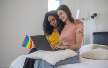 Lezbiyen çiftin yatakta dinlendiği hassas bir an birlikte tableti karıştırmayı seviyorlar. Konforlu yatak odası, samimi ve sıcak atmosfere katkı sağlıyor.