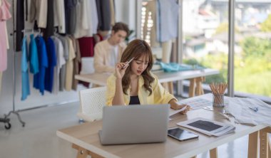 Moda tasarımcısı stüdyoda yeni kıyafet koleksiyonu için renk seçmeye odaklanmış durumda. Çalışma alanı yaratıcı araçlar, kumaş örnekleri, dikiş makinesi, canlı moda tasarım süreci ile dolu.