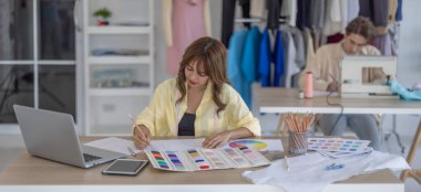 Moda tasarımcısı stüdyoda yeni kıyafet koleksiyonu için renk seçmeye odaklanmış durumda. Çalışma alanı yaratıcı araçlar, kumaş örnekleri, dikiş makinesi, canlı moda tasarım süreci ile dolu.