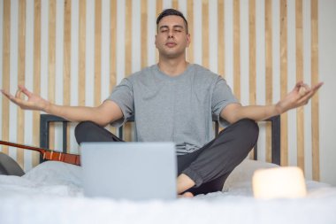 Sakin, loş bir yatak odasında, önünde dizüstü bilgisayarla meditasyon yapan bir adam ve yanında hafifçe parlayan bir mum, sakin ve odaklanmış bir atmosfer yaratıyor. Dijital detoks rutini