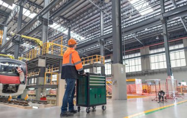 Demiryolu bakım mühendisi modern bir tren bakım tesisinde çalışıyor, bir alet arabasının yanında duruyor, güvenlik ve teknik hassasiyete odaklanıyor, diğer bir mühendis arka plandaki treni inceliyor.