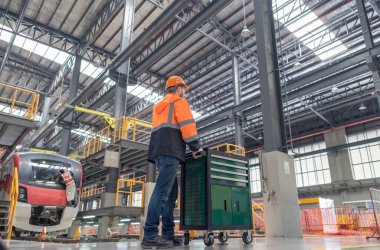 Demiryolu bakım mühendisi modern bir tren bakım tesisinde çalışıyor, bir alet arabasının yanında duruyor, güvenlik ve teknik hassasiyete odaklanıyor, diğer bir mühendis arka plandaki treni inceliyor.