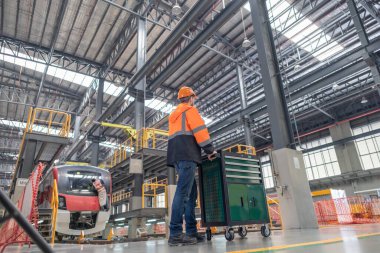 Demiryolu bakım mühendisi modern bir tren bakım tesisinde çalışıyor, bir alet arabasının yanında duruyor, güvenlik ve teknik hassasiyete odaklanıyor, diğer bir mühendis arka plandaki treni inceliyor.