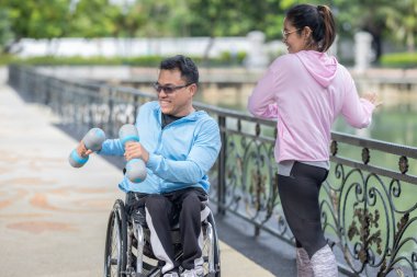 Tekerlekli sandalyede egzersiz yapan bir adam, bir direniş, güçlendirme ve kardiyo çalışması sırasında partneri tarafından destekleniyor. Bu resim fitness, motivasyon ve azim temalarını vurgular.