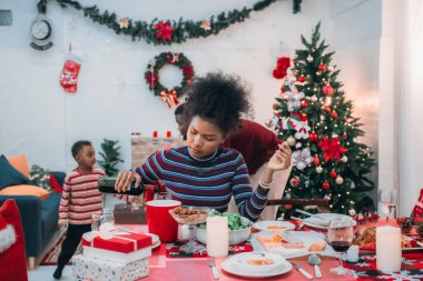 Siyah ailenin Noel yemeğinin keyfini çıkardığı canlı bir sahne, Noel mevsiminin sıcaklığını ve neşesini, güzel dekore edilmiş masaların etrafında yakalıyor, anne, baba ve oğulun özel anlarını paylaştığı.