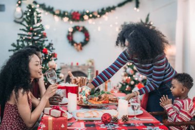 Sıcak ve neşeli bir Noel yemeği sahnesi. Siyah aile sohbete daldı ve birlikte güzel süslenmiş Noel ağacı ve tatil masasıyla süslenmiş bayram yemeğinin tadını çıkardı.