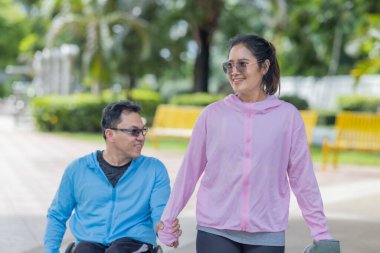 Parkta güneşli bir günün tadını çıkaran bir çift, tekerlekli sandalyedeki adam ve yanında yürüyen kadınla. Sahne, sakin açık hava ortamında arkadaşlığı, kapsayıcılığı ve aktif yaşam tarzını vurguluyor