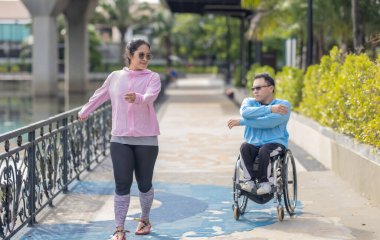 Parkta güneşli bir günün tadını çıkaran bir çift, tekerlekli sandalyedeki adam ve yanında yürüyen kadınla. Sahne, sakin açık hava ortamında arkadaşlığı, kapsayıcılığı ve aktif yaşam tarzını vurguluyor