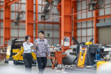 İki profesyonel, bir erkek ve bir kadın, yoğun bir metal işleme fabrikasında yürürken, takım çalışması ve sanayi uzmanlığını ön plana çıkararak işletim süreçlerini tartışıyor. CO2 emisyonları optimize ediliyor.