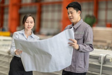 Bir erkek ve kadın mühendis, fabrika zeminindeki planları tartışıp gözden geçirir ve tüm özelliklerin metal levha fabrikasındaki üretim gereksinimleriyle aynı hizaya gelmesini sağlar..