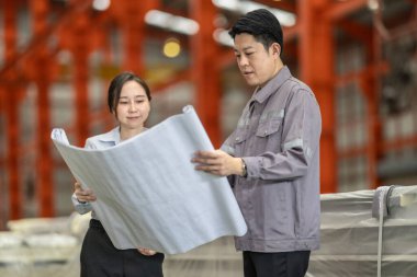 Bir erkek ve kadın mühendis, fabrika zeminindeki planları tartışıp gözden geçirir ve tüm özelliklerin metal levha fabrikasındaki üretim gereksinimleriyle aynı hizaya gelmesini sağlar..