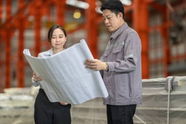 Bir erkek ve kadın mühendis, fabrika zeminindeki planları tartışıp gözden geçirir ve tüm özelliklerin metal levha fabrikasındaki üretim gereksinimleriyle aynı hizaya gelmesini sağlar..