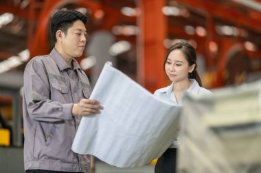 Bir erkek ve kadın mühendis, fabrika zeminindeki planları tartışıp gözden geçirir ve tüm özelliklerin metal levha fabrikasındaki üretim gereksinimleriyle aynı hizaya gelmesini sağlar..