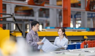 Bir erkek ve kadın mühendis, fabrika zeminindeki planları tartışıp gözden geçirir ve tüm özelliklerin metal levha fabrikasındaki üretim gereksinimleriyle aynı hizaya gelmesini sağlar..