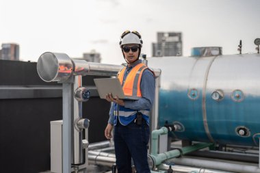 Profesyonel sanayi mühendisi, dizüstü bilgisayarla donatılmış, modern bir tesisin karmaşık boru hattı altyapısı içinde titizlikle bir HVAC sistemine sorun çıkarıyor. Karbon ayak izi azaltılıyor.