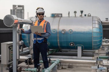 Profesyonel sanayi mühendisi, dizüstü bilgisayarla donatılmış, modern bir tesisin karmaşık boru hattı altyapısı içinde titizlikle bir HVAC sistemine sorun çıkarıyor. Karbon ayak izi azaltılıyor.