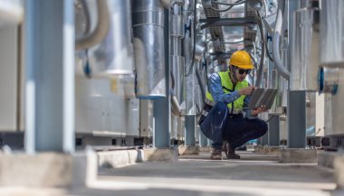 Profesyonel sanayi mühendisi, dizüstü bilgisayarla donatılmış, modern bir tesisin karmaşık boru hattı altyapısı içinde titizlikle bir HVAC sistemine sorun çıkarıyor. Karbon ayak izi azaltılıyor.