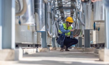 Profesyonel sanayi mühendisi, dizüstü bilgisayarla donatılmış, modern bir tesisin karmaşık boru hattı altyapısı içinde titizlikle bir HVAC sistemine sorun çıkarıyor. Karbon ayak izi azaltılıyor.