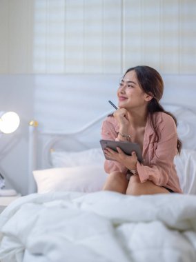 Genç bir kadın rahat yatak odasında tablet kullanırken yaratıcı düşüncelere daldı. Bir ilham ve üretkenlik anını resmediyordu.