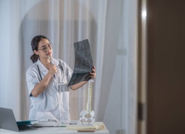 Bir kadın tıp ve ortopedi öğrencisi, iyi aydınlatılmış bir çalışma odasında, anatomik modeller ve ders kitaplarıyla çevrili MR filmlerini incelikle inceleyerek bir öğrenme ve keşif anı sergiliyor..