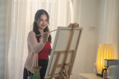 Genç bir kadın kendi sanat eserlerine dikkatle odaklanıyor, kendi stüdyosundaki bir sehpaya kurulmuş bir tuval üzerine resim yapıyor. Sahne rahat ve yaratıcı bir atmosfer yakalıyor. Odada yumuşak doğal ışık süzüyor.