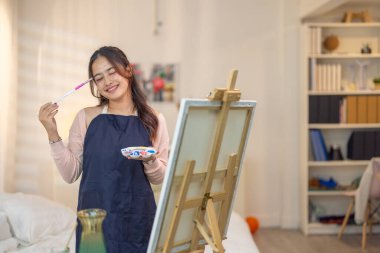 Genç bir kadın kendi sanat eserlerine dikkatle odaklanıyor, kendi stüdyosundaki bir sehpaya kurulmuş bir tuval üzerine resim yapıyor. Sahne rahat ve yaratıcı bir atmosfer yakalıyor. Odada yumuşak doğal ışık süzüyor.