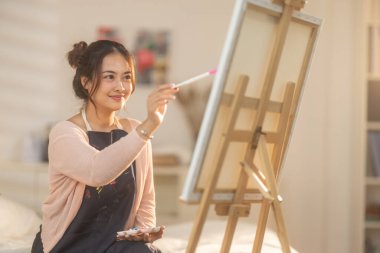 Genç bir kadın kendi sanat eserlerine dikkatle odaklanıyor, kendi stüdyosundaki bir sehpaya kurulmuş bir tuval üzerine resim yapıyor. Sahne rahat ve yaratıcı bir atmosfer yakalıyor. Odada yumuşak doğal ışık süzüyor.