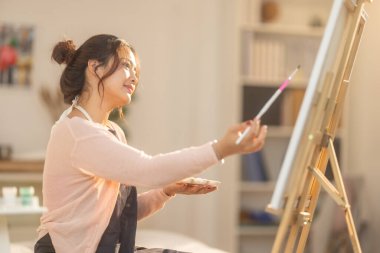 Genç bir kadın kendi sanat eserlerine dikkatle odaklanıyor, kendi stüdyosundaki bir sehpaya kurulmuş bir tuval üzerine resim yapıyor. Sahne rahat ve yaratıcı bir atmosfer yakalıyor. Odada yumuşak doğal ışık süzüyor.