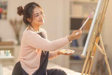 Genç bir kadın kendi sanat eserlerine dikkatle odaklanıyor, kendi stüdyosundaki bir sehpaya kurulmuş bir tuval üzerine resim yapıyor. Sahne rahat ve yaratıcı bir atmosfer yakalıyor. Odada yumuşak doğal ışık süzüyor.