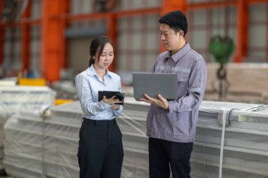 Metal fabrikasındaki stratejik planlama oturumunda çalışan bir erkek ve kadın arasında dijital aletlerin iş akışını artırdığı odaklı bir etkileşim.