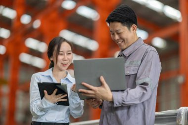 Metal fabrikasındaki stratejik planlama oturumunda çalışan bir erkek ve kadın arasında dijital aletlerin iş akışını artırdığı odaklı bir etkileşim.