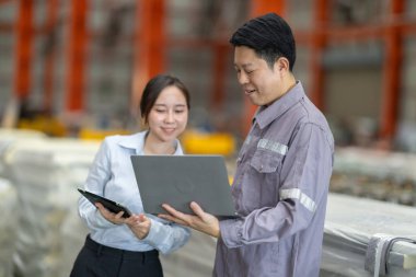 Metal fabrikasındaki stratejik planlama oturumunda çalışan bir erkek ve kadın arasında dijital aletlerin iş akışını artırdığı odaklı bir etkileşim.