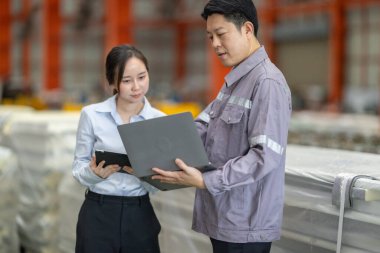 Metal fabrikasındaki stratejik planlama oturumunda çalışan bir erkek ve kadın arasında dijital aletlerin iş akışını artırdığı odaklı bir etkileşim.