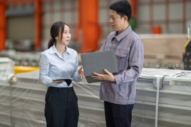 Metal fabrikasındaki stratejik planlama oturumunda çalışan bir erkek ve kadın arasında dijital aletlerin iş akışını artırdığı odaklı bir etkileşim.