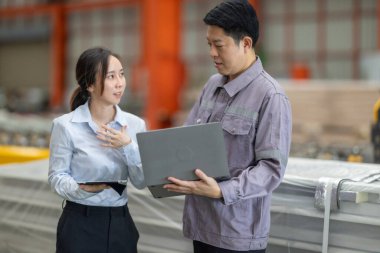 Metal fabrikasındaki stratejik planlama oturumunda çalışan bir erkek ve kadın arasında dijital aletlerin iş akışını artırdığı odaklı bir etkileşim.