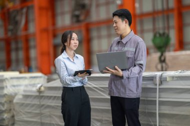 Metal fabrikasındaki stratejik planlama oturumunda çalışan bir erkek ve kadın arasında dijital aletlerin iş akışını artırdığı odaklı bir etkileşim.