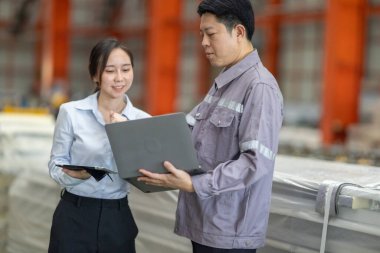 Metal fabrikasındaki stratejik planlama oturumunda çalışan bir erkek ve kadın arasında dijital aletlerin iş akışını artırdığı odaklı bir etkileşim.