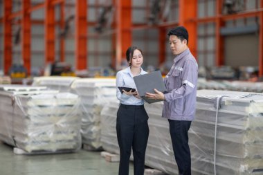 Metal fabrikasındaki stratejik planlama oturumunda çalışan bir erkek ve kadın arasında dijital aletlerin iş akışını artırdığı odaklı bir etkileşim.