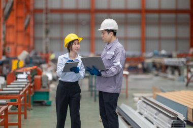 Metal fabrikasındaki stratejik planlama oturumunda çalışan bir erkek ve kadın arasında dijital aletlerin iş akışını artırdığı odaklı bir etkileşim.