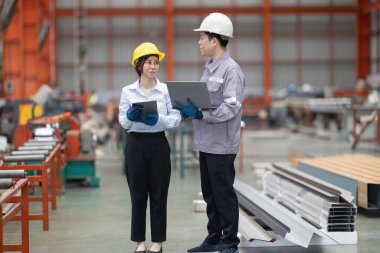 Metal fabrikasındaki stratejik planlama oturumunda çalışan bir erkek ve kadın arasında dijital aletlerin iş akışını artırdığı odaklı bir etkileşim.