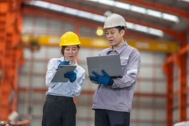 Metal fabrikasındaki stratejik planlama oturumunda çalışan bir erkek ve kadın arasında dijital aletlerin iş akışını artırdığı odaklı bir etkileşim.