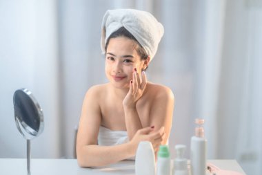 Saçına havlu sarılmış neşeli bir kadın gösteriş için otururken yüz kremi sürüyor. Cilt bakımı ürünleri ve ayna görünür olup, rahatlatıcı ve sıcak ortamlarda kişisel bakım rutinini vurgular.